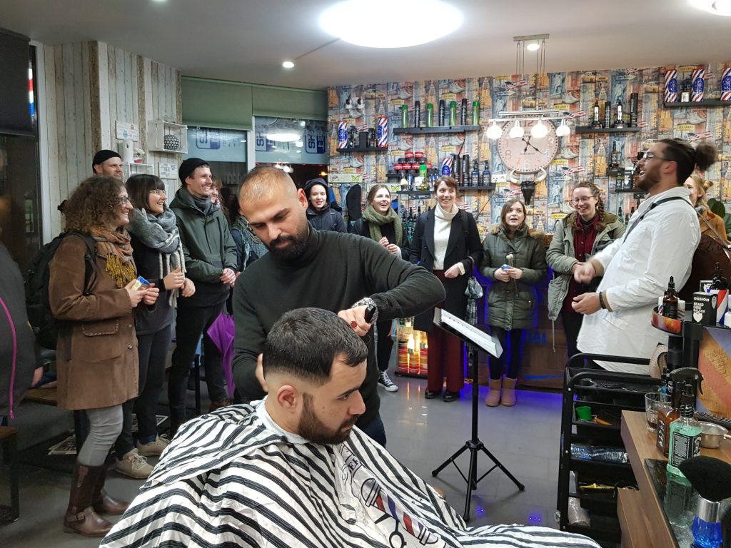 Im Vordergrund werden einem Herrn die Haare geschnitten, im Hintergrund im Friseursalon stehen Menschen und singen