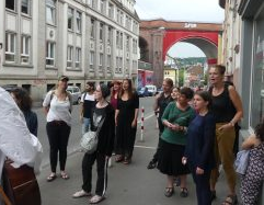 Eine Gruppe Menschen singt an einer befahrenen Straße, im Hintergrund ein hohes Viadukt