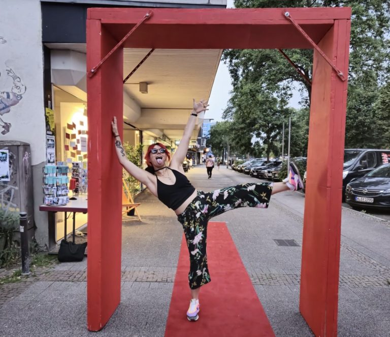 Eine Frau steht unter einem roten Bogen auf einem roten Teppich und streckt ein Bein gerade in die Luft