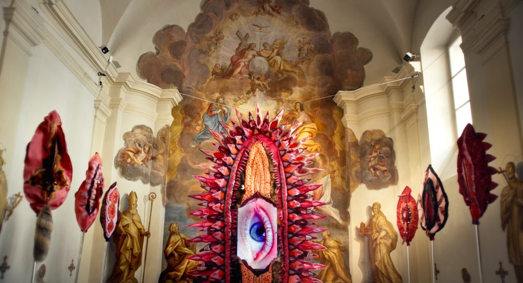 Künstlerisch gestaltete Vulven stehen in einer Kirche vor einem Wandgemälde