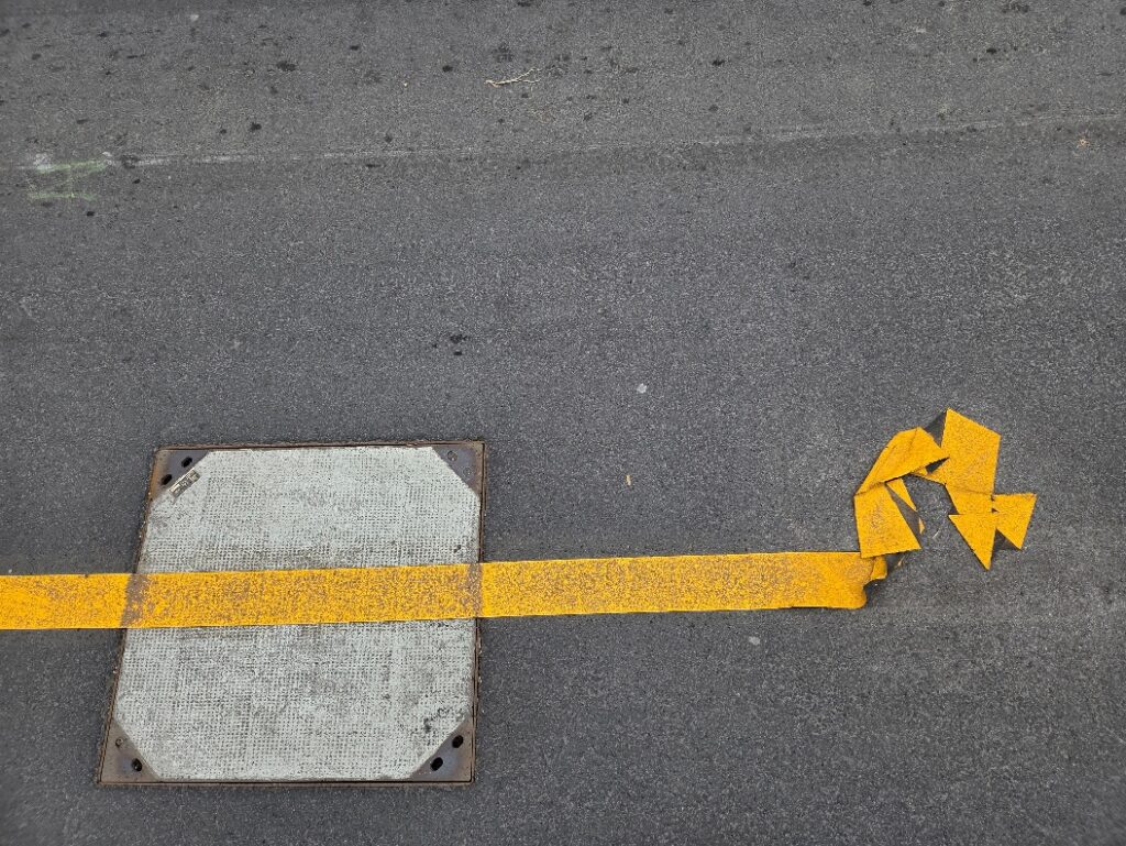 Straße mit Kanaldeckel, daruf ein Steifen von einer Straßenmarkierung
